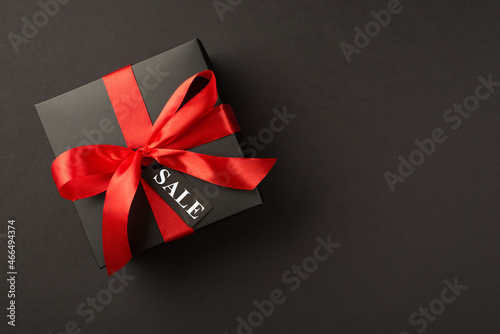 Top view photo of stylish giftbox in black packaging with vivid red satin ribbon bow and tag on isolated black background with text sale on pricetag
