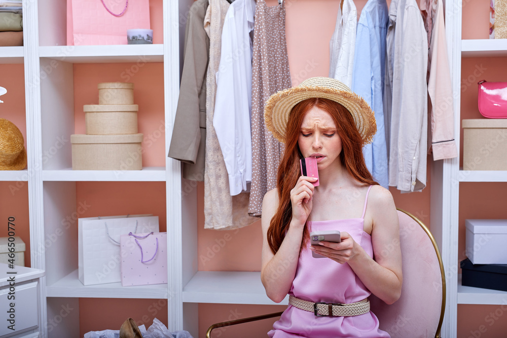 Obraz premium nervous red-haired woman using shopping apps on smartphone, doing online shopping, sitting among lots of colourful trendy clothes in wardrobe. attractive caucasian lady in pink dress at home