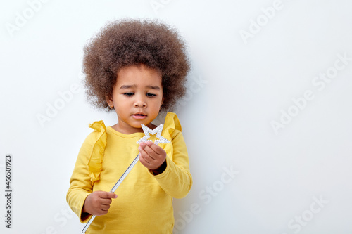 Pretty cute afro american child girl with fluffy curly hair holding magic wand in hands looking at it with interest, wondering, looking curious. isolated on white studio background, portrait © alfa27