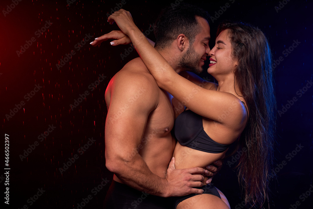 Naklejka premium smiling young man gently hugging wet sensual woman in water drops on black background, portrait of caucasian lovely couple with sportive athletic body posing at camera, on love. in rain
