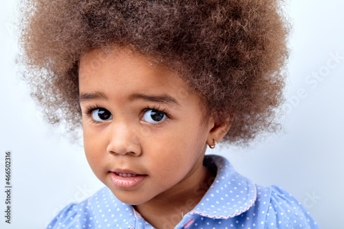 Cute multiracial small girl with funny expression look at camera isolated on white background. Confused child is looking at something, having charming look, fluffy curly hair. copy space