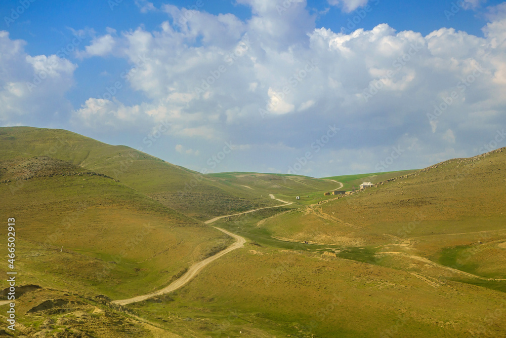 Country road winding between hills of valley. Village houses and small figures of residents are visible near hill on right side. Shot in Qashqadaryo region of Uzbekistan