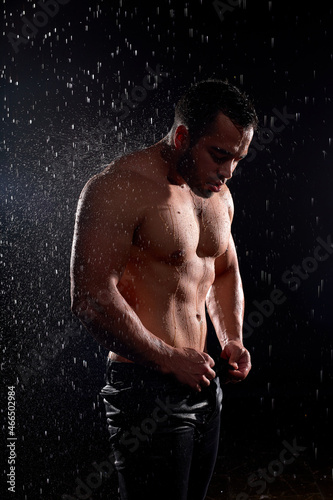 Powerful sexy male model with Muscular body, athlete with big muscles posing at camera, isolated on black background, in rain, water drops on body. Side view portrait. Bodybuilding, sport, people