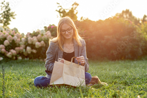 Hungy Caucasian businesswoman, student or freelancer with craft paper bag siting in the park, ordering takeaway food delivery. Concept of rest and break on job. photo