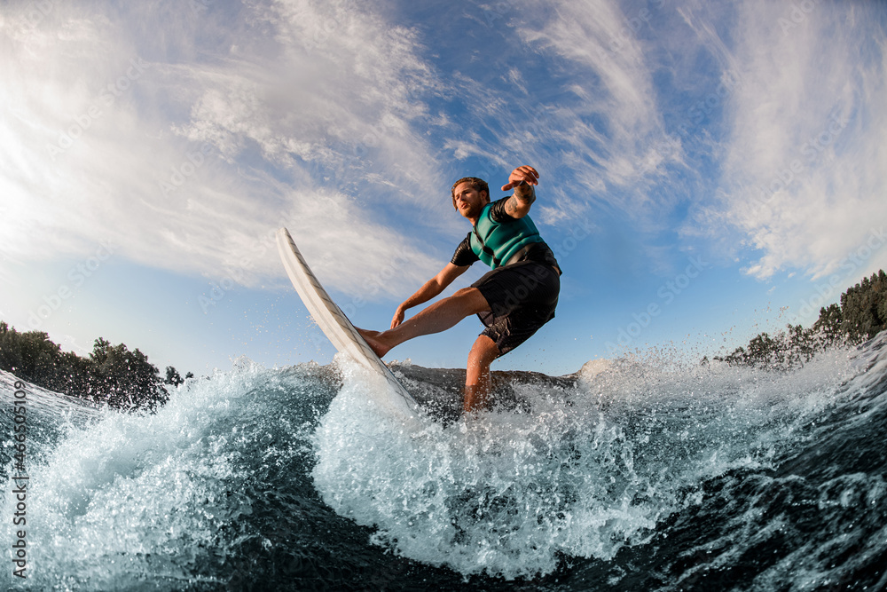 athletic man wakesurfer masterfully riding down the blue splashing wave on a warm day