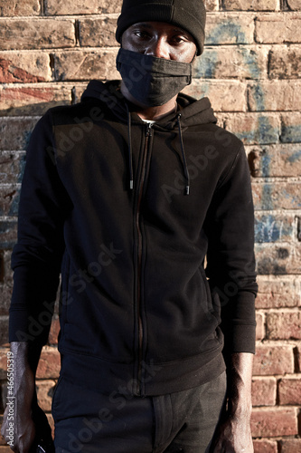 Portrait of young african man in mask standing by brick wall, hooligan in black wear. black guy pose a danger for society, waiting for victim, commit a crime. social problems, crime concept