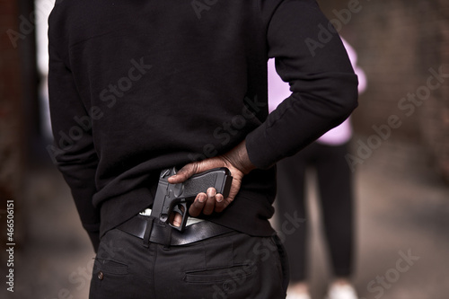 Robbery or criminal man in black hoodie is holding gun standing behind the woman  going to commit a crime. view from back on male in black outfit  close-up hands. criminal activity concept