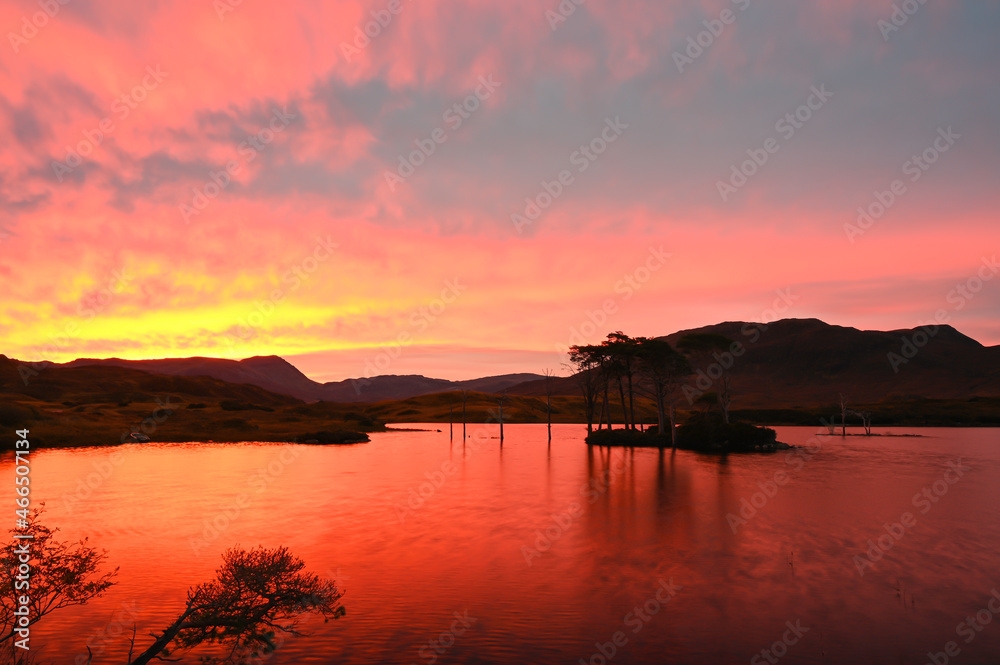 Stunning sunrise over Loch Assynt in Sutherland, Scottish Highlands. On NC500 route. Silhouettes of trees on island on loch, hills in background. Red, orange, yellow and blue sky reflected in water.