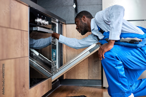 focused african man or service worker in special clothing installs, disassembles or performs maintenance of dishwasher built into kitchen furniture, side view on black guy in blue overalls photo