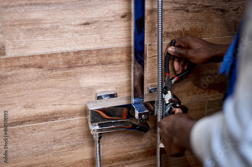 Installing shower faucet. Black Man's hands fixing shower faucet to the place. using worktool pliers, close-up photo. Side view. Plumbing, repairing, assemble concept. Cropped male in blue overalls photo