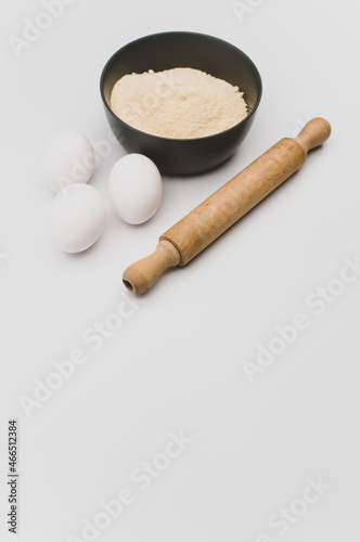 dark stoneware bowl with flour wooden rolling pin and white eggs on a white tabletop