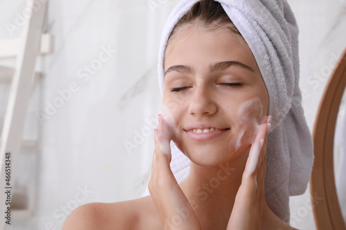 Beautiful teenage girl applying cleansing foam onto face in bathroom, space for text. Skin care cosmetic