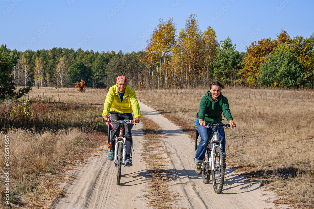 Fototapeta premium Man and woman ride bicycles on a country road through a golden birch grove in autumn