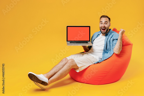 Full body young fun man in blue shirt white t-shirt sit in bag chair hold use work on laptop pc computer with blank screen workspace area do winner gesture isolated on plain yellow background studio.