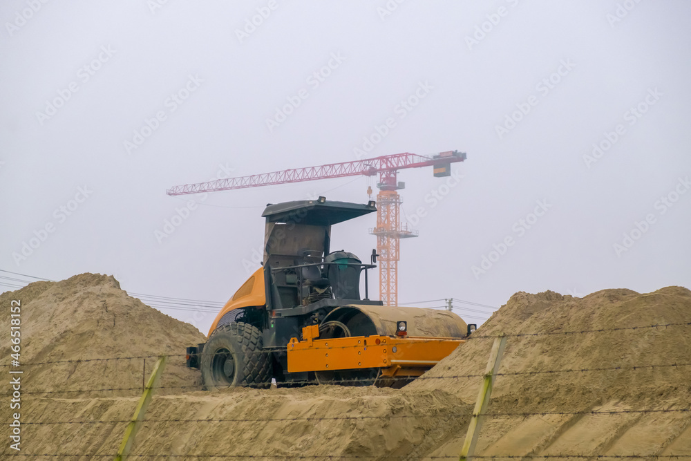 Obraz premium Yellow road compactor on a construction site. Construction equipment on the background of an object under construction. Tower cranes on a construction site.