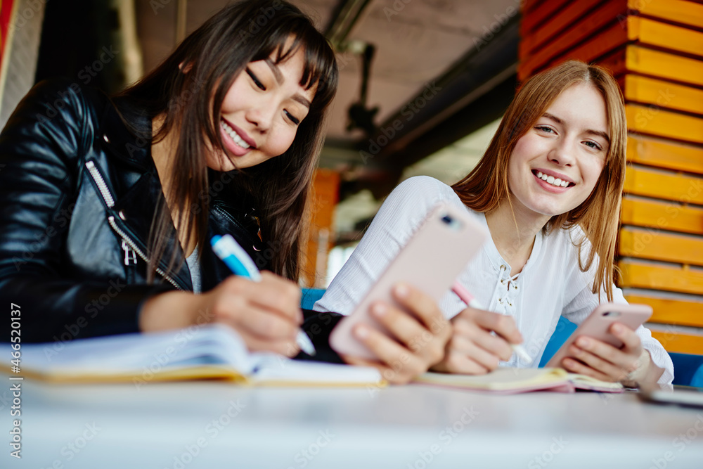 Portrait Caucasian female student smiling at camera while Asian friend making informative notes in textbook, cheerful women bloggers with cellphone gadget posing and studying in university Portrait Caucasian female student smiling at camera while Asian friend making informative notes in textbook, cheerful women bloggers with cellphone gadget posing and studying in university