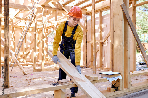 confident female constructor working on wooden construction site, wearing working uniform clothes, in gloves and hardhat, replacing wooden beam into another place, alone, outdoors.