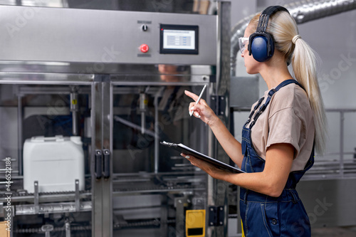 skilled engineer woman rate the quality of pesticides production in factory, take notes and think. professional female industrier holding folde-tablet and pen in hands, wearing uniform and headset