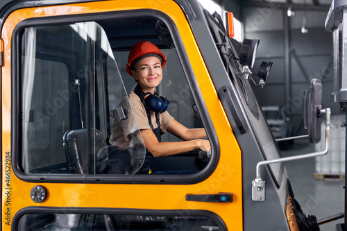 Beautiful woman forklift worker operator driving vehicle at warehouse, factory. female driving loader confidently, feminism and male work concept. lady in safety hardhat and uniform