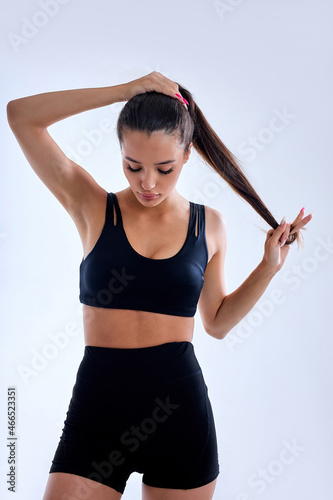 Image of muscular young fit woman posing in sportswear against white background. Caucasian female model with perfect torso in studio, making ponytail, lady is looking down. portrait