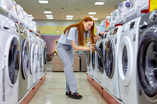 Woman buying new clothes washer in supermarket, choosing the best and the most modern. Redhead Lady In Casual Wear Standing Between Various Different Washine Machines, Make Choice and purchase photo