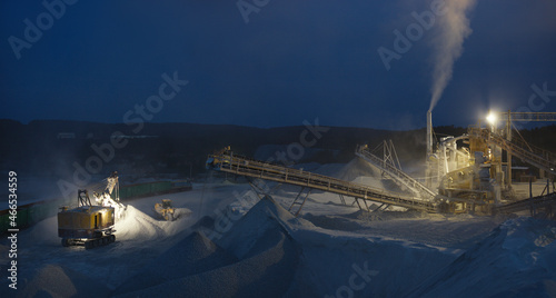 Excavator loads crushed stone into railway wagons near to the metal structures of stone crushing equipment at a mining plant, night panorama with artificial lighting.