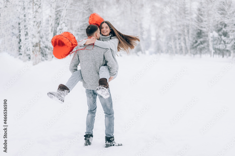A man holds a woman in his arms in front of a snow park. A woman holds in her hands heart-shaped balloons filled with helium