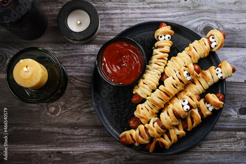 Cute Halloween party snack: wiener sausage with sugar eyes wrapped in dough stripes resembling mummies on a black plate with tomato ketchup