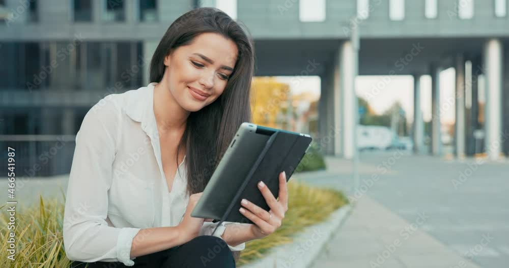 Beautiful ambitious businesswoman in white shirt sits in front of glass modern building of insurance corporation company, woman holds tablet in hand, makes video call, waves to camera, break from work Beautiful ambitious businesswoman in white shirt sits in front of glass modern building of insurance corporation company, woman holds tablet in hand, makes video call, waves to camera, break from work