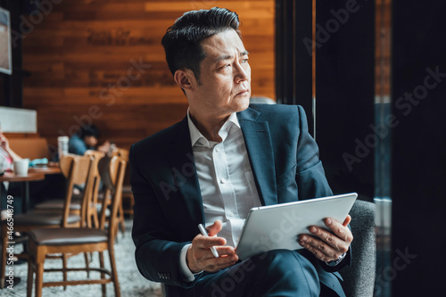 Handsome Asian Businessman Using Digital Tablet in a Cafe. Serious business man in blue suit working on a digital tablet while sitting in a restaurant and looking away.