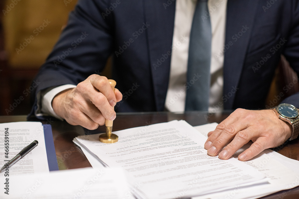Hands of director in elegant suit putting stamp on approved document such as certificate, contract, application form or permit paper Hands of director in elegant suit putting stamp on approved document such as certificate, contract, application form or permit paper