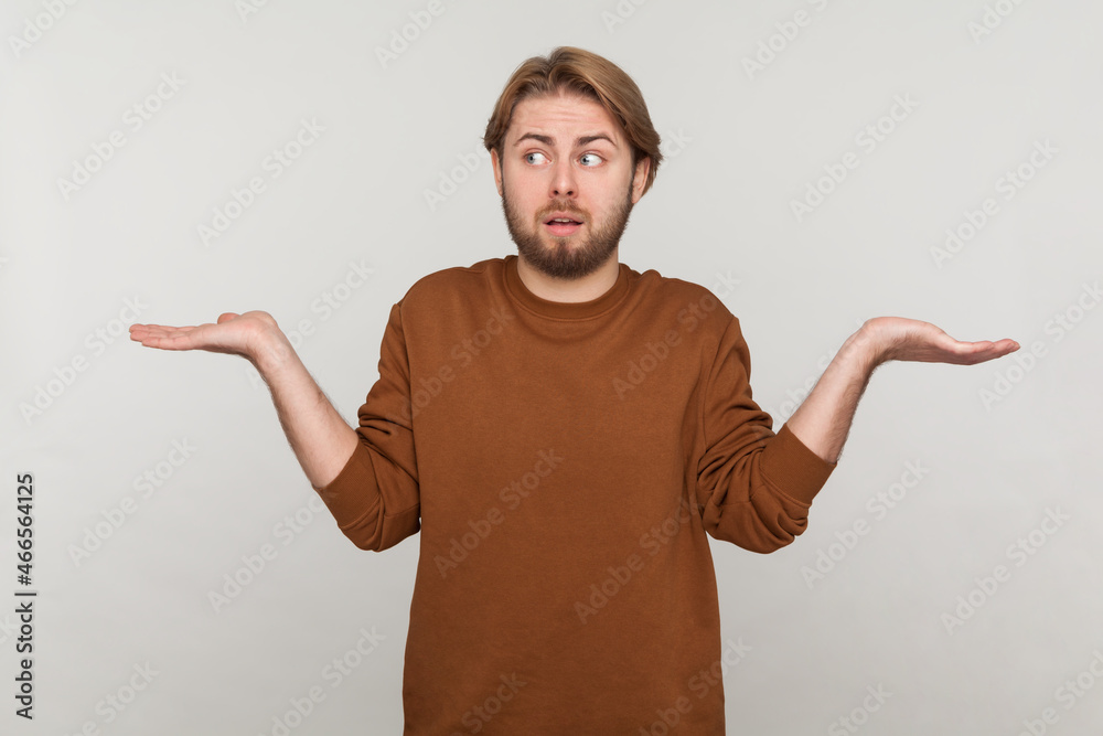 Portrait of man with beard wearing sweatshirt, shrugging shoulders as doesn't know answer, can`t make decision, being uncertain, not sure. Indoor studio shot isolated on gray background.
