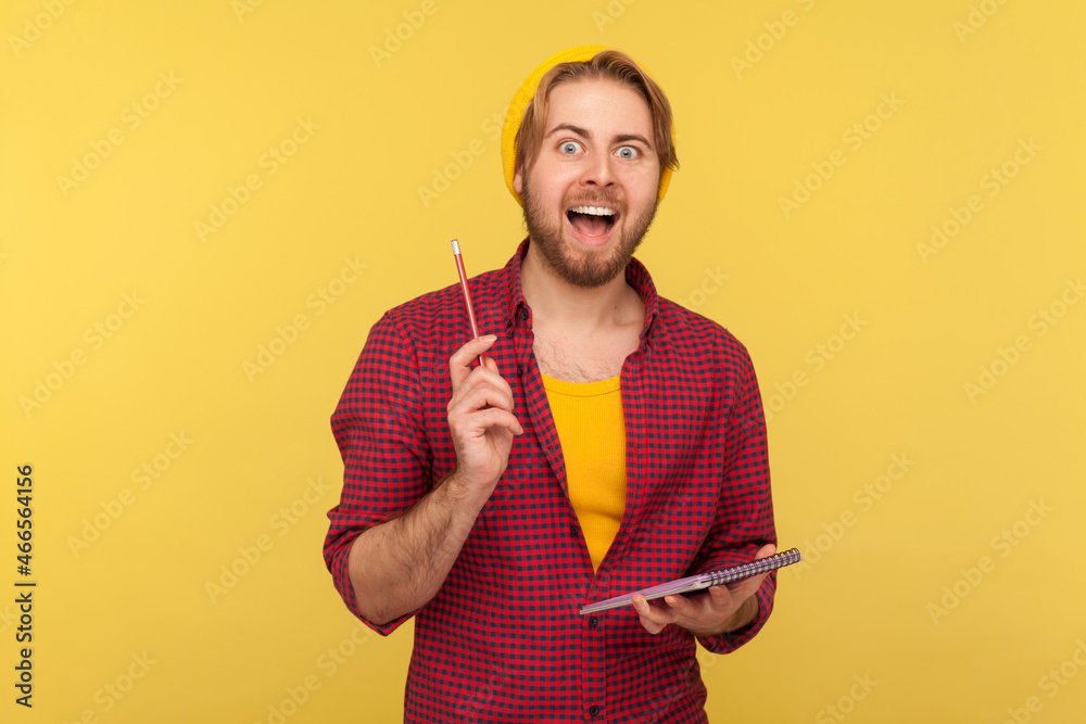 Portrait of hipster bearded guy in beanie hat and checkered shirt writing creative idea into notebook, drawing sketch or taking notes in paper. Indoor studio shot isolated on yellow background. Portrait of hipster bearded guy in beanie hat and checkered shirt writing creative idea into notebook, drawing sketch or taking notes in paper. Indoor studio shot isolated on yellow background.