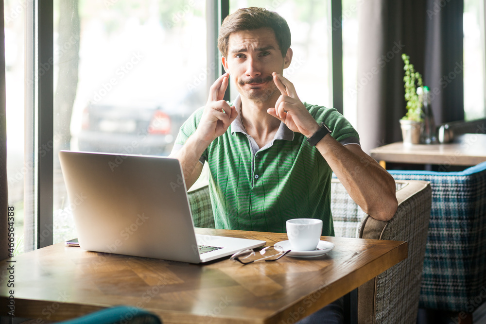 Portrait of man employee wearing green T-shirt, crossing fingers and wishing for good luck, in anticipation of success, working online on laptop, Indoor shot near big window, cafe background. Portrait of man employee wearing green T-shirt, crossing fingers and wishing for good luck, in anticipation of success, working online on laptop, Indoor shot near big window, cafe background.