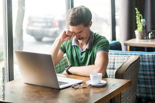 Portrait of sad businessman wearing green T-shirt, rubbing tired eyes, crying because of hopelessness and loneliness, nervous breakdown. Indoor shot near big window, cafe background.