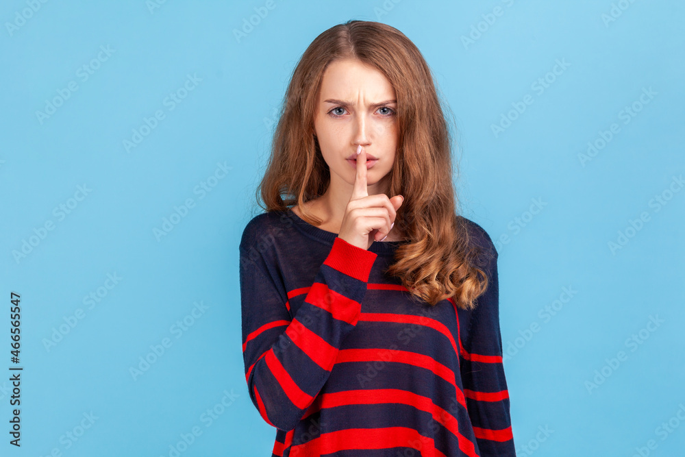 Keep silence and be quiet! Woman wearing striped casual style sweater looking angrily and holding finger on lips, gesturing shut up, stop talking. Indoor studio shot isolated on blue background. Keep silence and be quiet! Woman wearing striped casual style sweater looking angrily and holding finger on lips, gesturing shut up, stop talking. Indoor studio shot isolated on blue background.