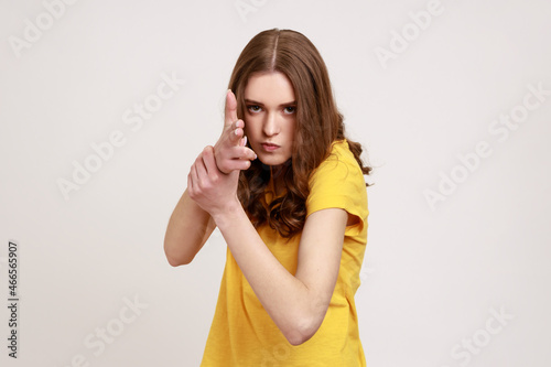 I'll kill you! Portrait of angry teenager girl in yellow T-shirt pointing finger guns to camera, threatening to shoot, hands imitating weapon. Indoor studio shot isolated on gray background.