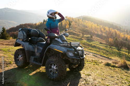 Young happy female driver in protective helmet enjoying extreme ride on ATV quad motorbike in fall mountains at sunset.