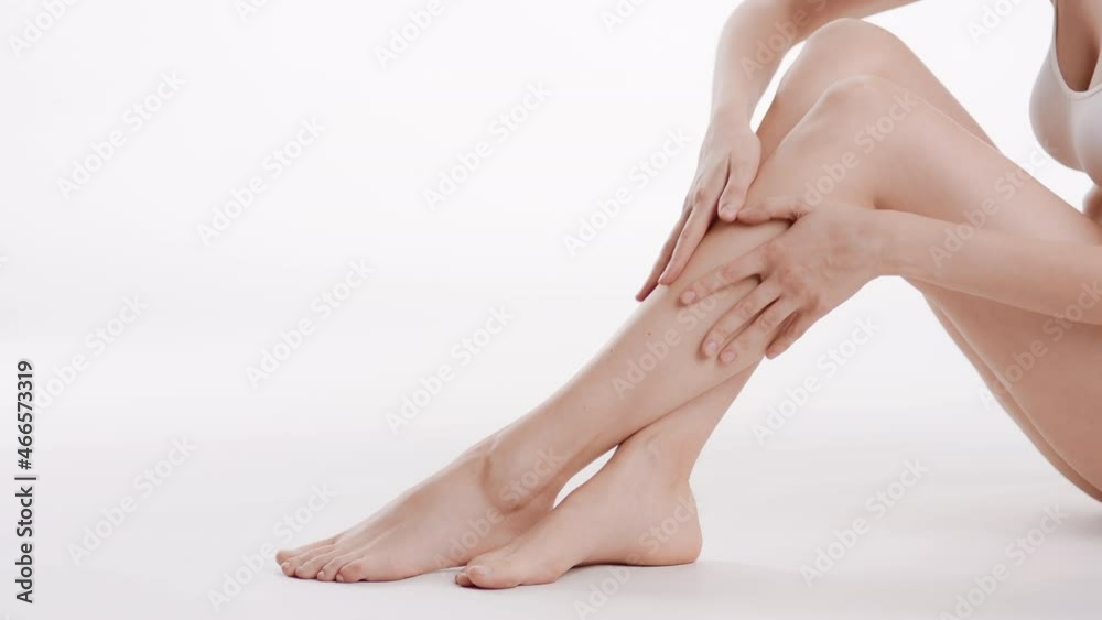 Good-looking slim female skincare model in white bra touches her crossed smooth legs sitting on the floor on white background | Body care concept
