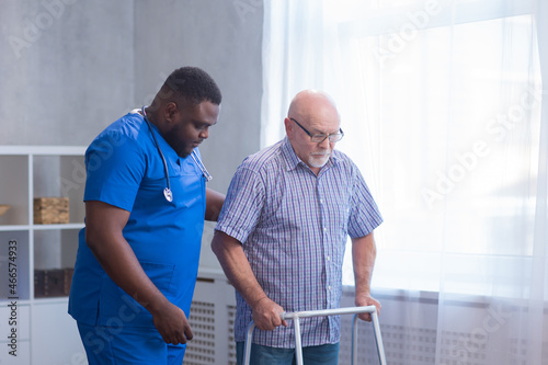 Caregiver is teaching old man to walk with walker. Professional nurse and patient in a nursing home. Assistance, rehabilitation and health.