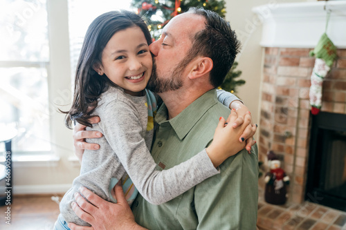 Dad hugging daughter and kissing her cheek in front of Christmas tree 
