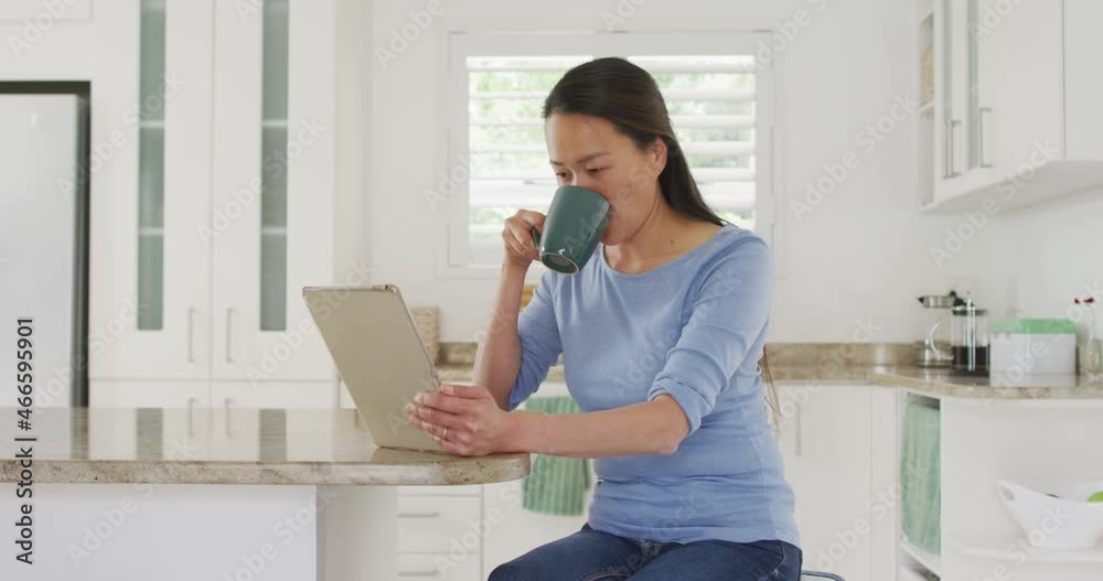 Happy asian woman sitting at table, drinking coffee and using tablet in kitchen Happy asian woman sitting at table, drinking coffee and using tablet in kitchen