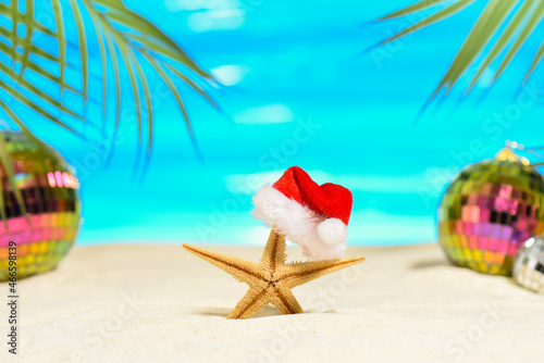 Tropical Merry Christmas concept. Starfishe in a Santa hat on the sandy seashore. Mirrored trending disco balls and palm branches in the background.