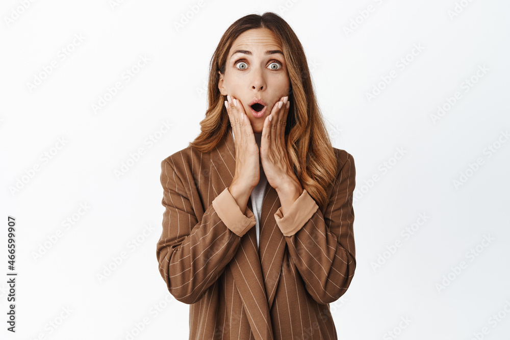 Portrate of surprise woman in suit looking at camera, say wow, touching her face and watching smth amazing, standing in awe against white background Portrate of surprise woman in suit looking at camera, say wow, touching her face and watching smth amazing, standing in awe against white background