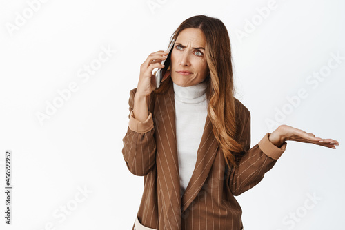 Fototapeta Confused businesswoman talking on cellphone, shrugging while listening caller, s