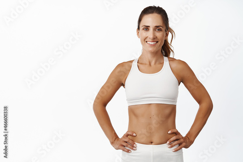 Confident sportswoman in white sportsbra, holding hands on waist, fitness trainer standing in power pose, workout in gym, white background