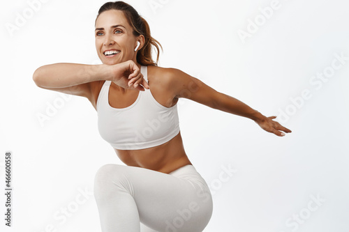 Portrait of sportswoman stretching body, exercising, doing fitness leg raise and smiling, running, standing over white background