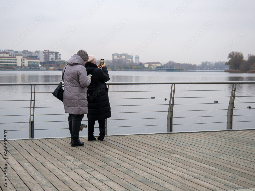 Women take pictures of the lake with ducks on their phones. Cloudy, autumn day in Russia.