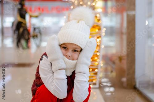 surprised boy in white bobble hat and scarf at mall near showcase. blurred bright lights on background
