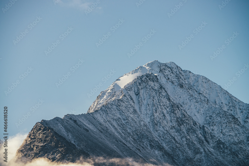 Minimalist sunny mountain landscape with snow-covered mountain top in gold sunshine in clear blue sky. Minimal alpine scenery with high snowy pinnacle in sunlight above low clouds. Snow on sharp rocks